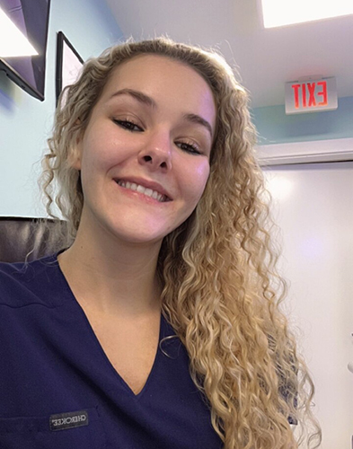 Young woman in a navy blue scrub top, smiling, with long curly blonde hair.